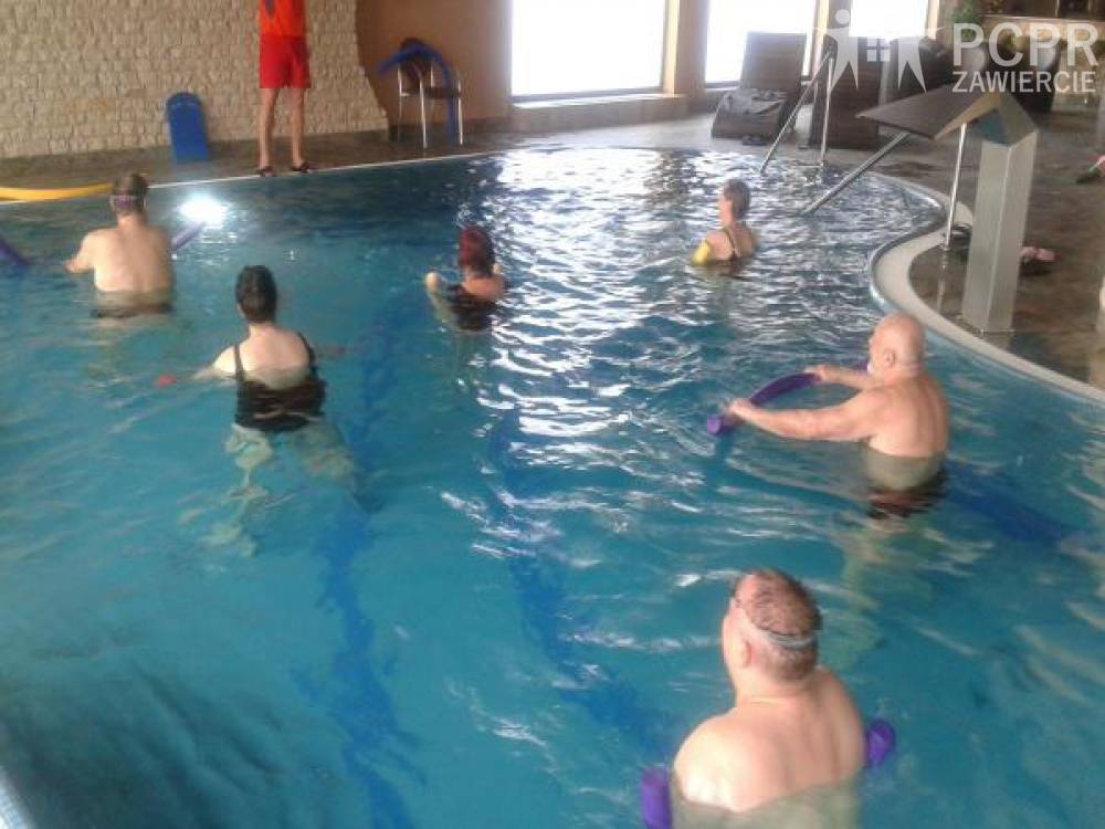 Zdjęcie: Grupa kobiet i mężczyzn stojąc w basenie wykonuje ćwiczenie trzymając piankowy makaron